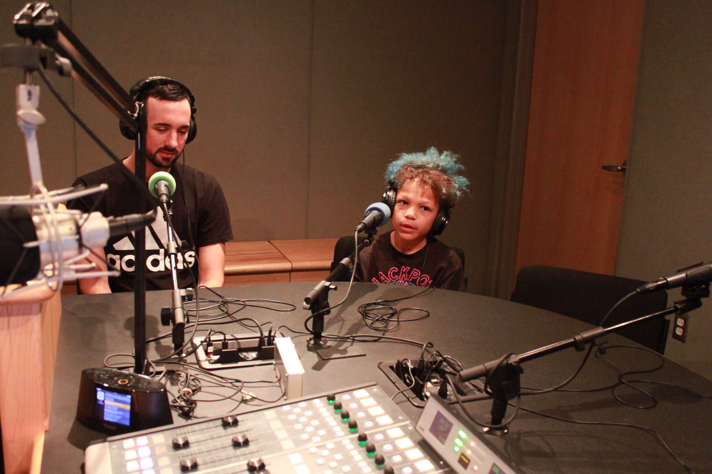 An adult and a child wearing headphones and speaking into microphones in a radio studio
