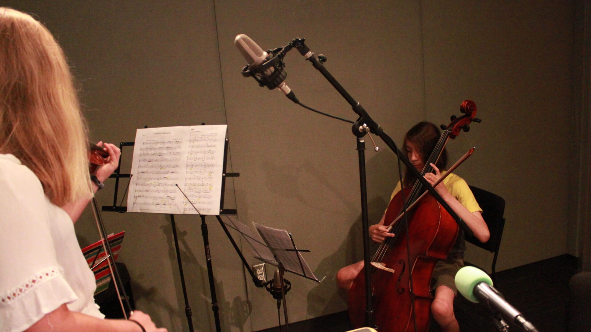 Youth playing cello and an individual playing a violin, both looking at sheet music on music stands in a radio studio