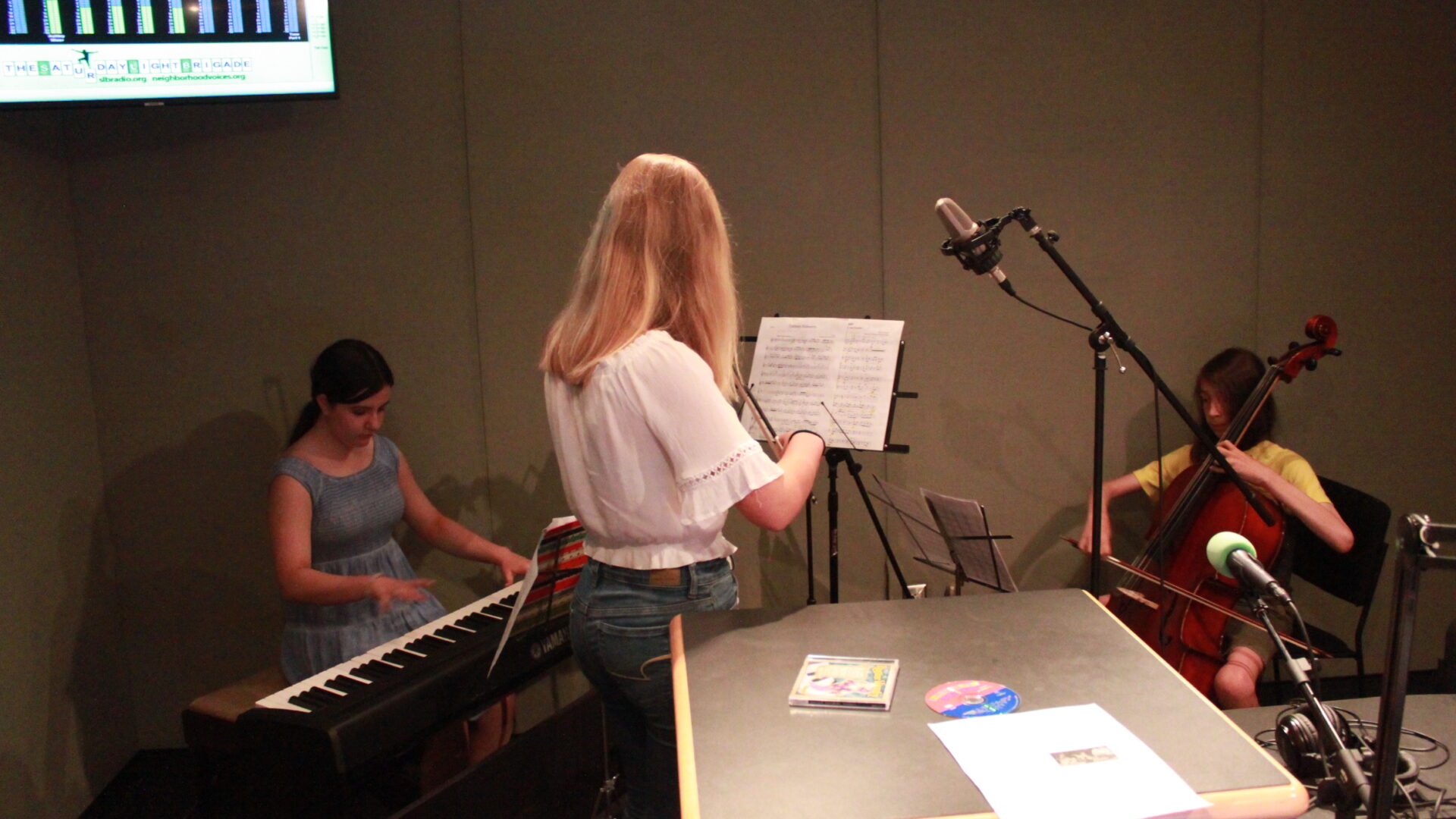 Three youth performing on a piano keyboard, a violin, and a cello looking at sheet music on music stands in a radio studio