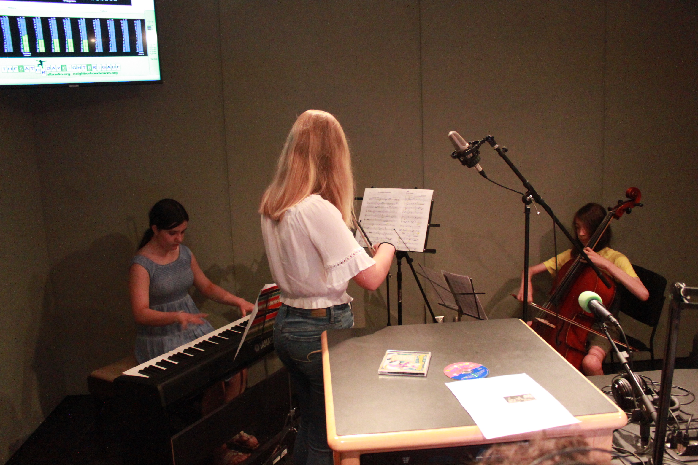 Three youth performing on a piano keyboard, a violin, and a cello looking at sheet music on music stands in a radio studio
