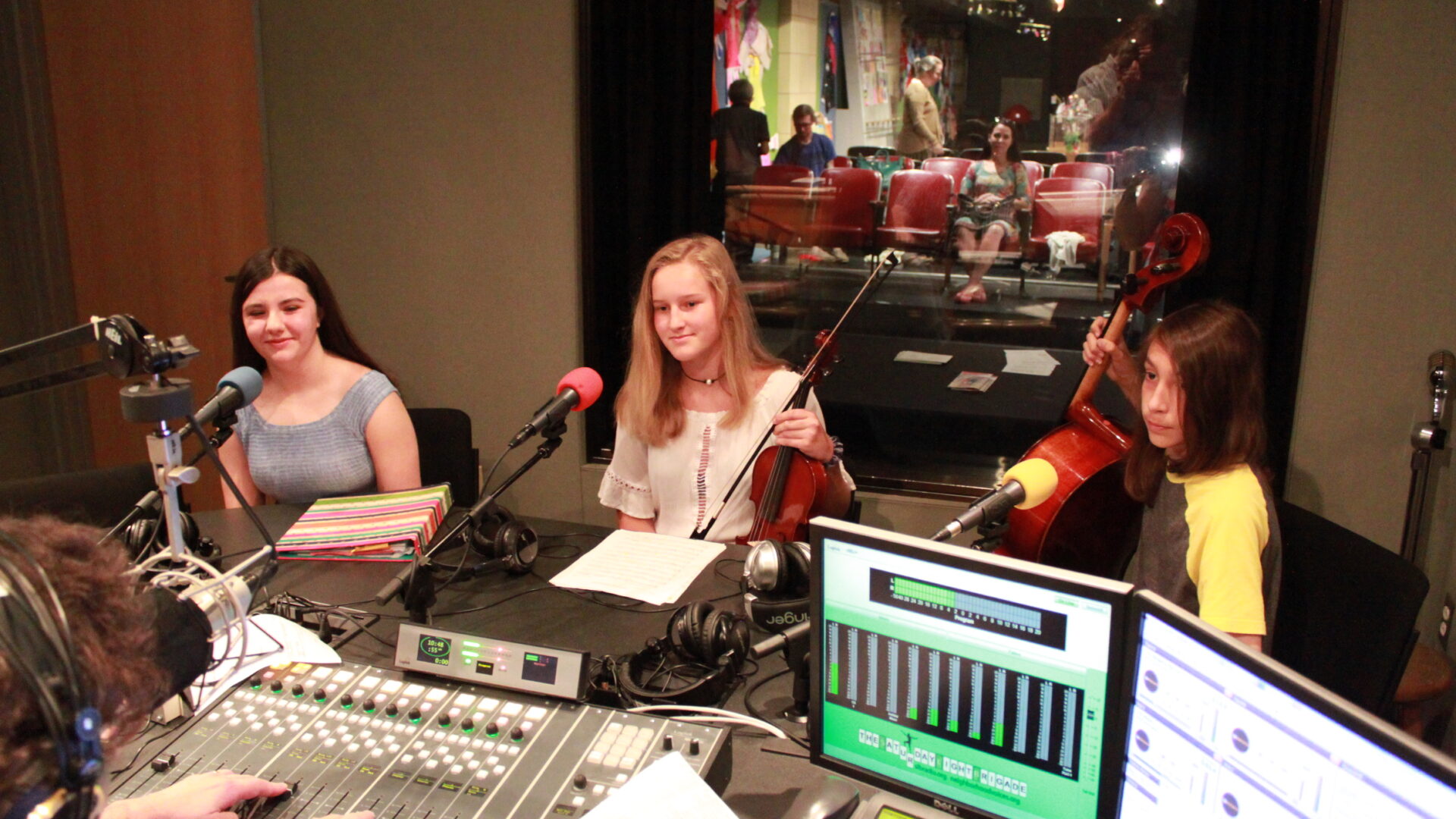 Three youth sitting in front of microphones being interviewed by an adult wearing headphones interacting with a mixing board in a radio studio, one youth holding a violin, one holding a cello