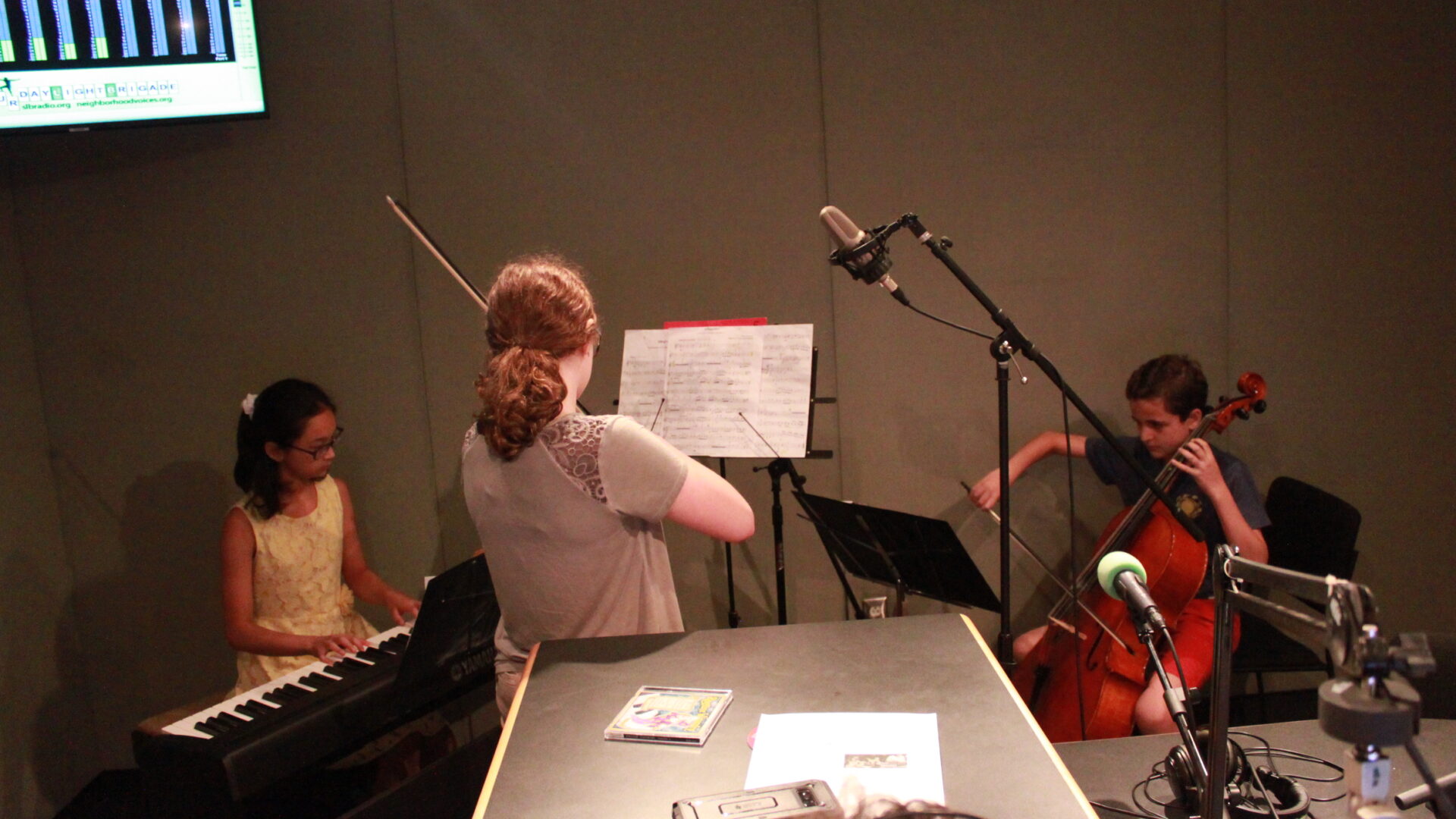 Three youth performing on piano keyboard, violin, and cello looking at sheet music on music stands in a radio studio