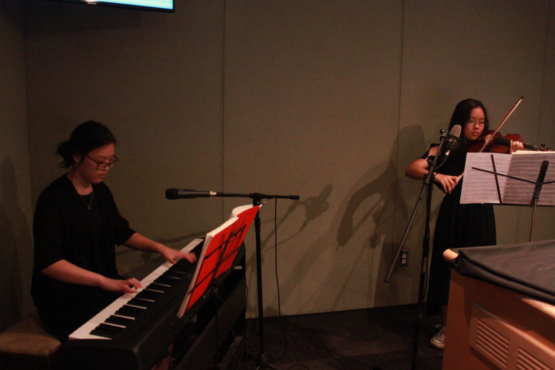 Two youth performing on piano keyboard and violin looking at sheet music on music stands in a radio studio