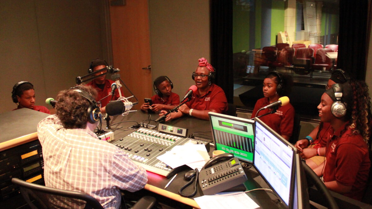 Six youth and one adult, all wearing headphones and speaking into microphones in a radio studio