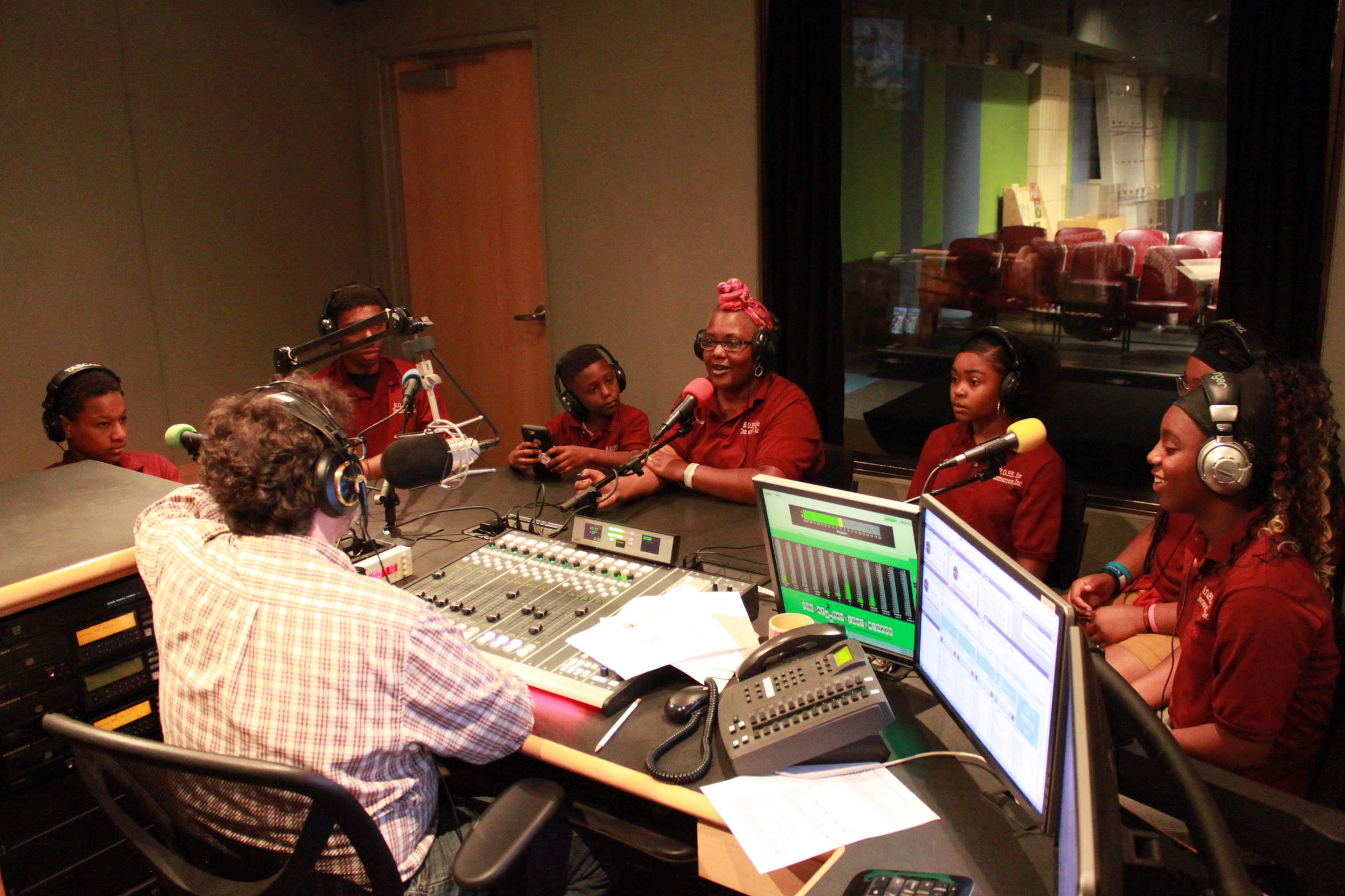 Six youth and one adult, all wearing headphones and speaking into microphones in a radio studio