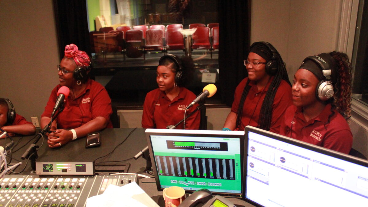 Three teens and one adult, all wearing headphones and speaking into microphones in a radio studio