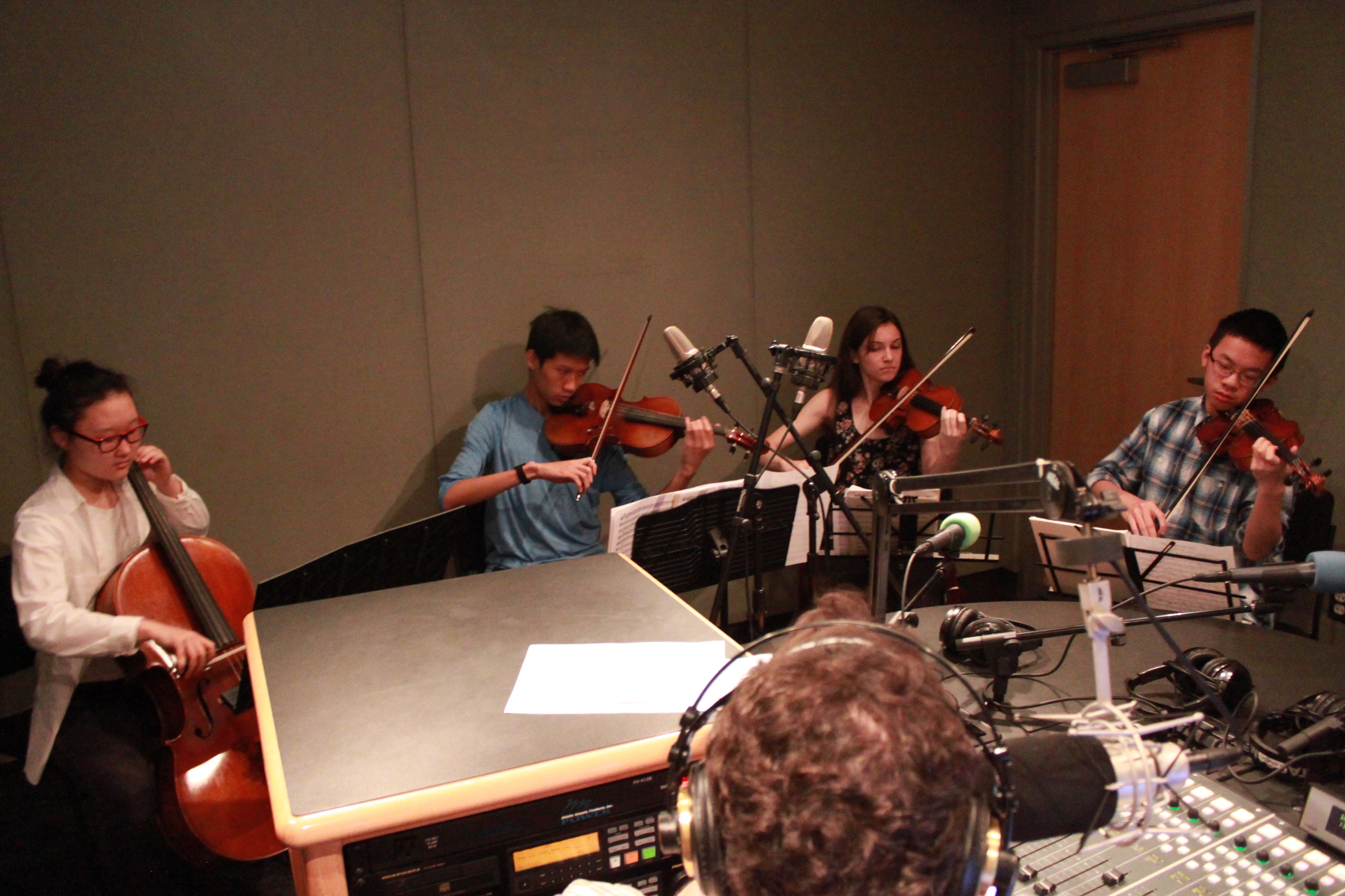 Four teen musicians performing on violin, viola, and cello looking at sheet music on music stands while an adult wearing headphones listens in a radio studio