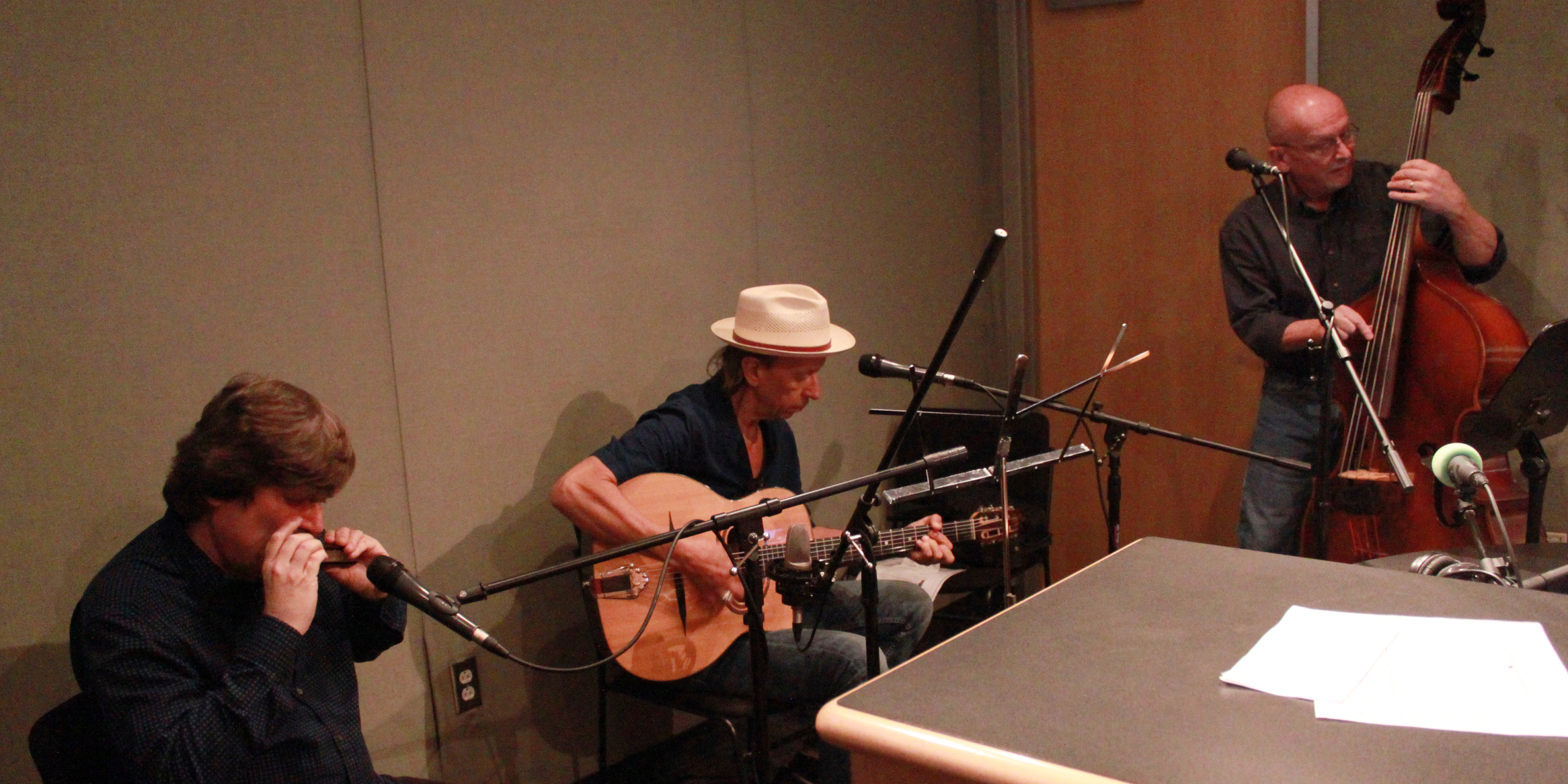 Three adult musicians performing on harmonica, guitar, and upright bass in a radio studio