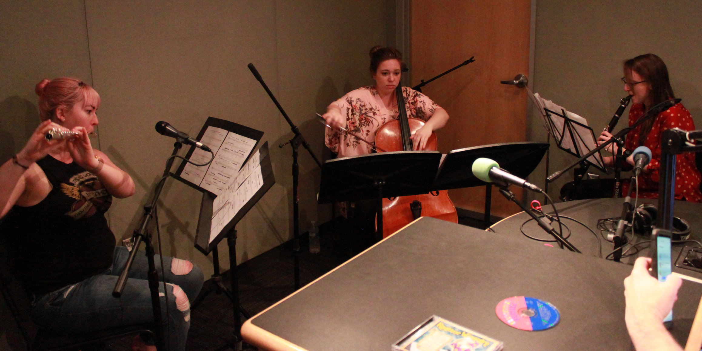 Three young adults performing on clarinet, flute, and cello looking at sheet music on music stands in a radio studio