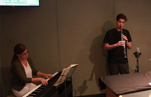 Teen playing clarinet into a microphone and adult playing a piano keyboard and looking at sheet music on a music stand in a radio studio