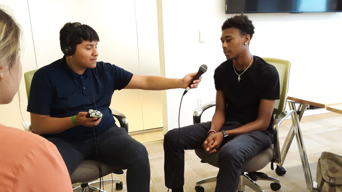 Teen wearing headphones and holding a portable recorder and a microphone interviewing another teen speaking into the microphone