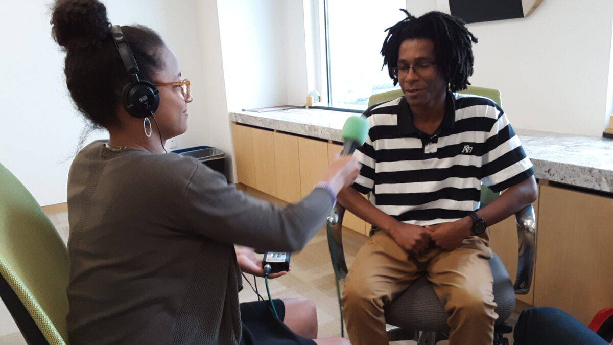 A teen wearing headphones and holding a portable recorder and a microphone interviewing another teen sitting down