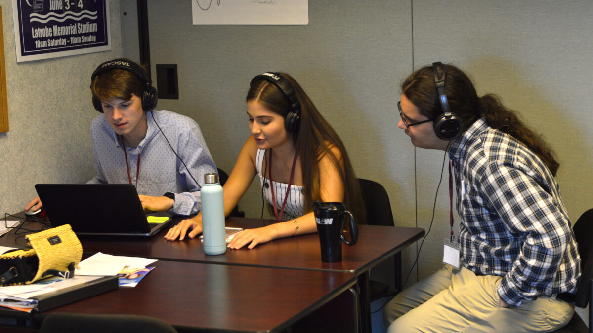 Three teens wearing headphones, one looking at a laptop, two looking at a smartphone