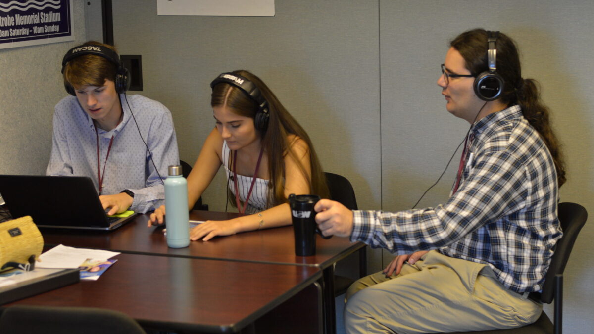 Three teens wearing headphones, one looking at a laptop, one looking at a smartphone, one holding a mug