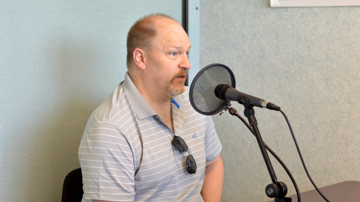 Adult with sunglasses hanging from shirt speaking into a microphone with a pop filter