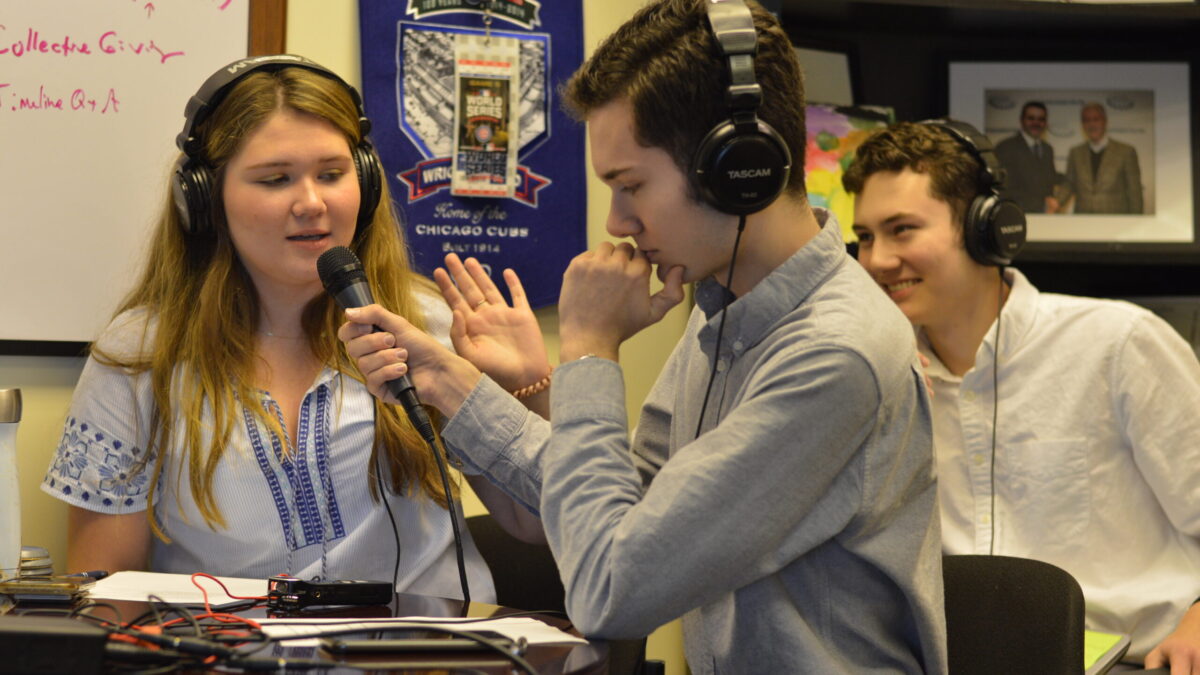Two teens wearing headphones interviewing another teen wearing headphones and speaking into a microphone connected to a portable recorder