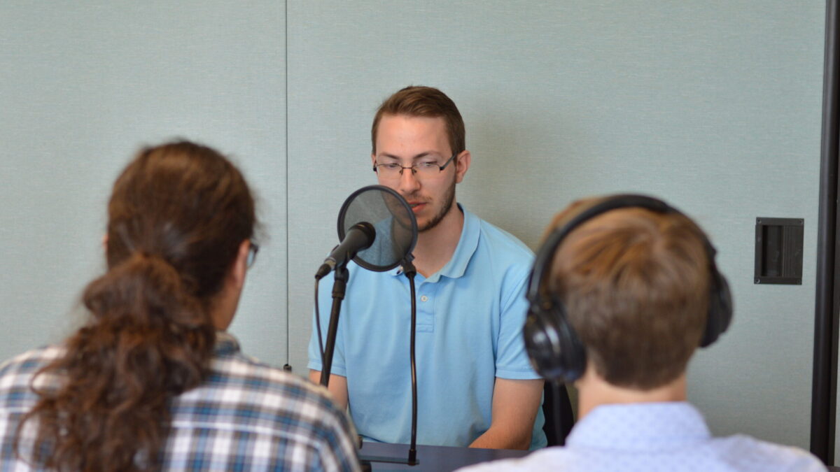 Two teens, one wearing headphones interviewing another teen speaking into a microphone with a pop filter