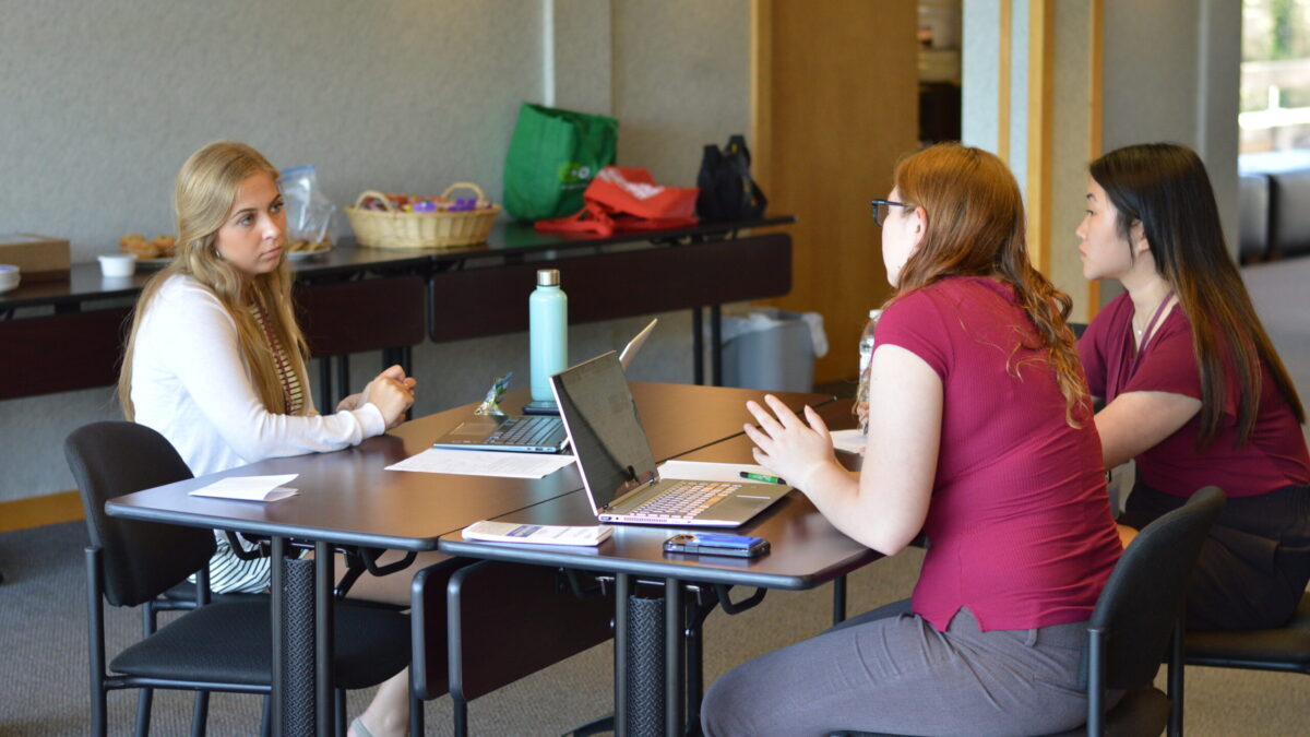 Three young adults having a discussion in front of open laptops on two black tables in a gray room