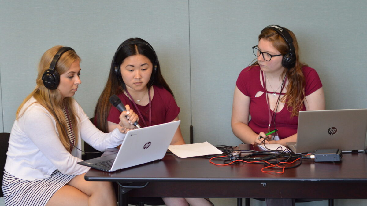 Two teens wearing headphones and one adult wearing headphones and holding a microphone looking at a laptop computer on a wooden table