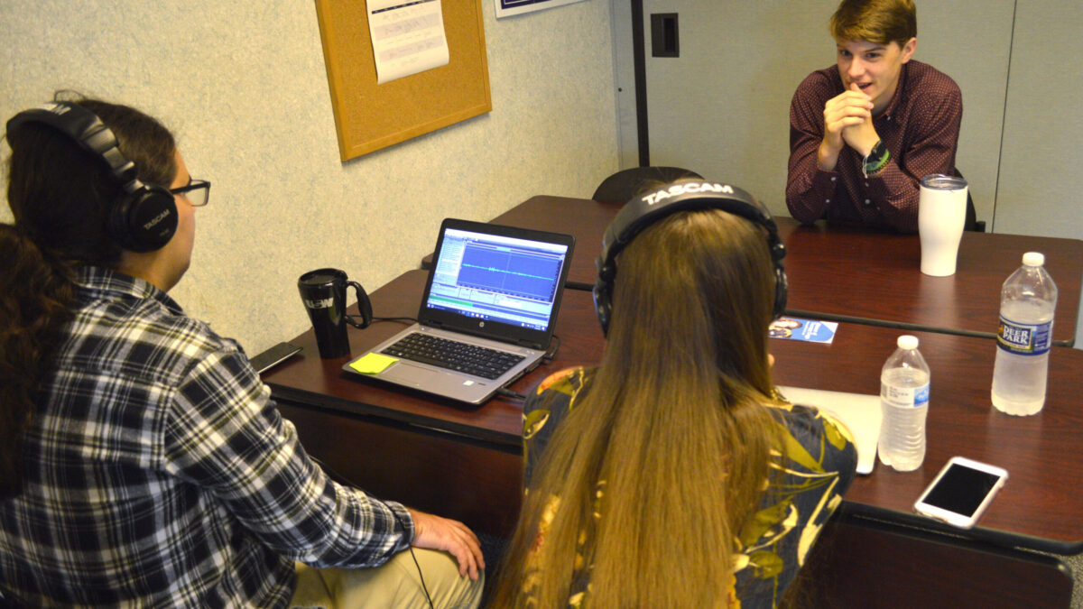Two teens wearing headphones looking at a laptop computer sitting across from another teen