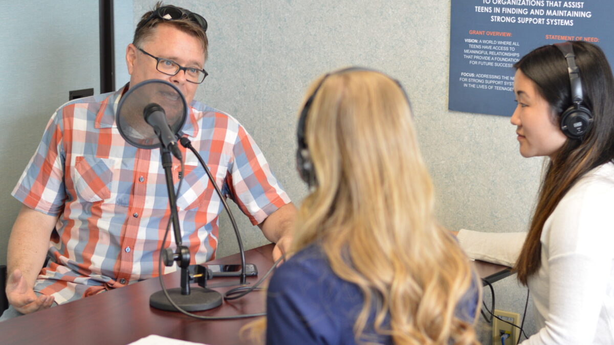 Two teens wearing headphones interviewing an adult speaking into a microphone sitting in front of a smartphone on a wooden table