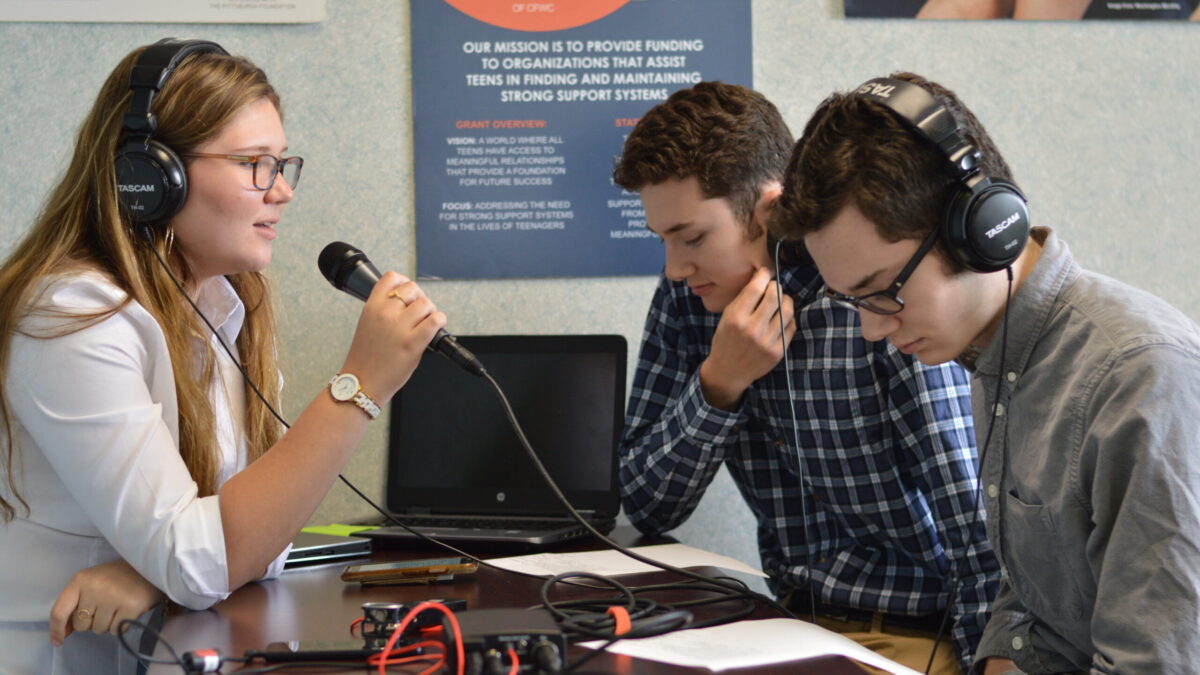 Two teens wearing headphones interviewing another teen wearing headphones and holding a microphone