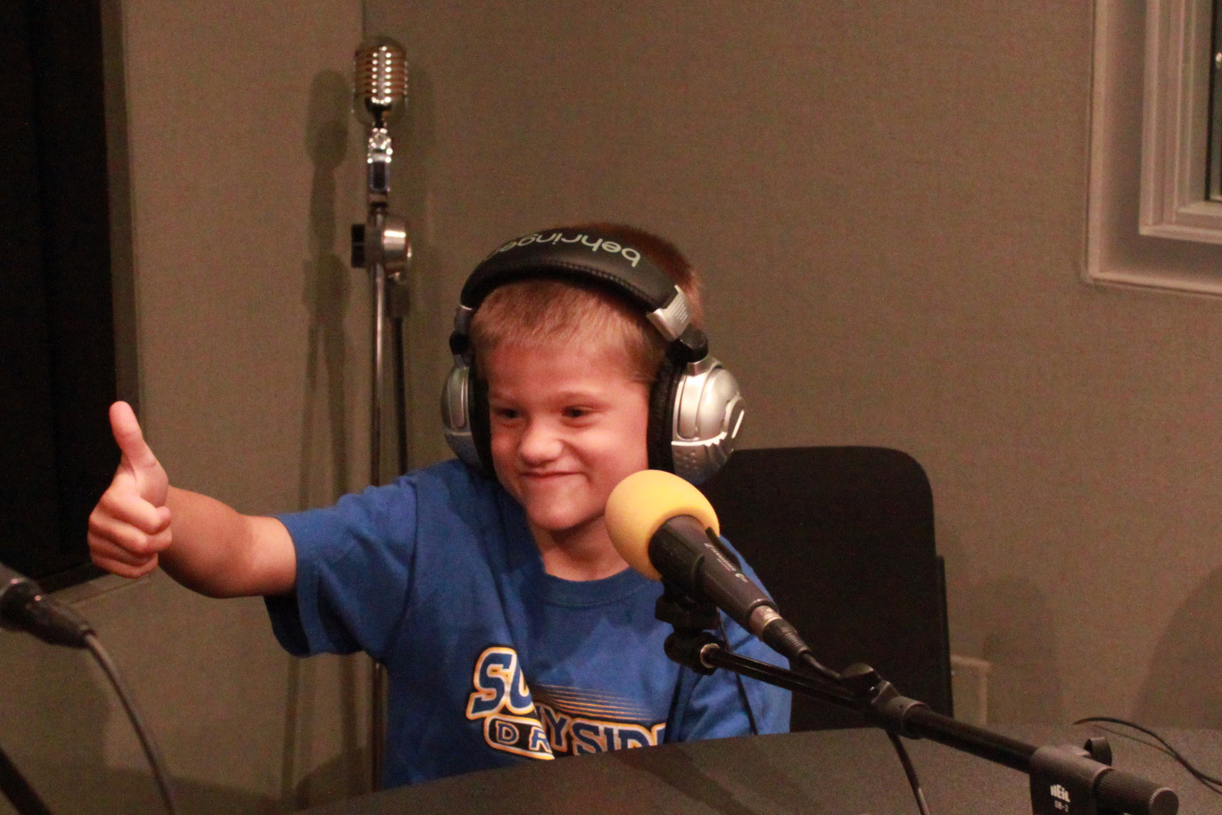 Child wearing headphones sitting in front of a microphone giving a thumbs up in a radio studio