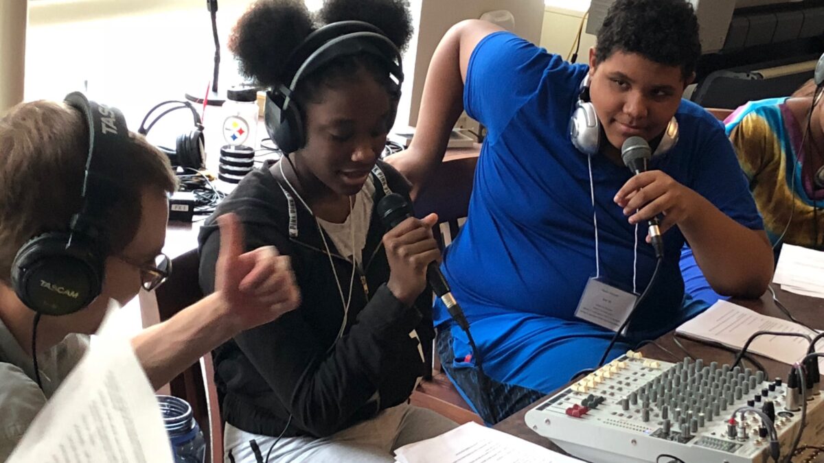 Three teens wearing headphones, two talking into microphones, one giving the other two a thumbs up