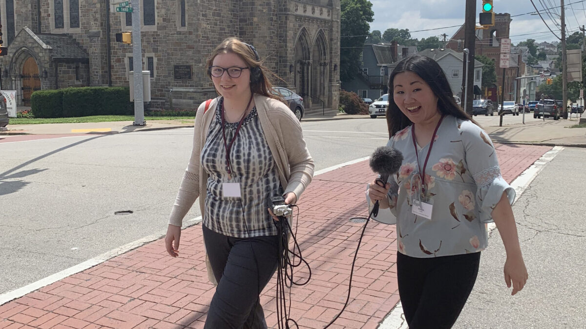 Two teens smiling at the camera while walking on a crosswalk, one wearing headphones and holding a portable recorder, the other holding a microphone