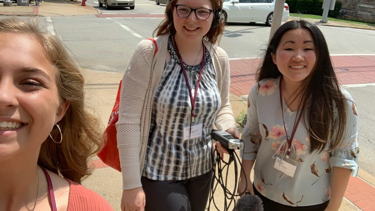 Three teens smiling at the camera, one wearing headphones and holding a portable recorder, one holding a microphone