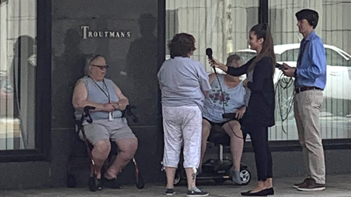 A teen wearing headphones and holding a portable recorder and a teen holding a microphone interviewing three adults on the sidewalk