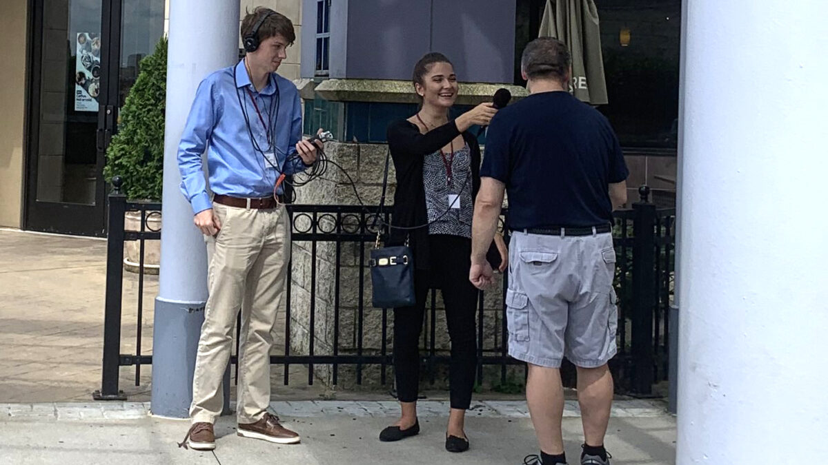 A teen wearing headphones and holding a portable recorder and another teen holding a microphone interviewing an adult on the sidewalk