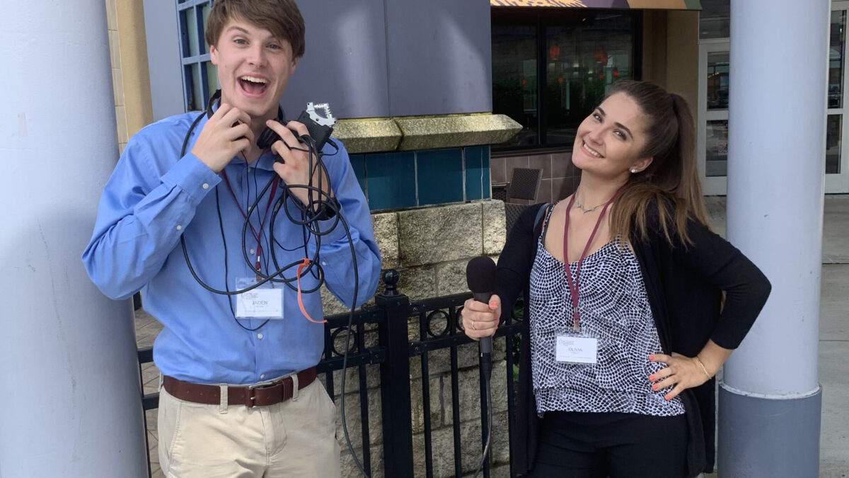 A teen holding headphones around their neck and a portable recorder and another teen holing a microphone, both smiling and posing in profile