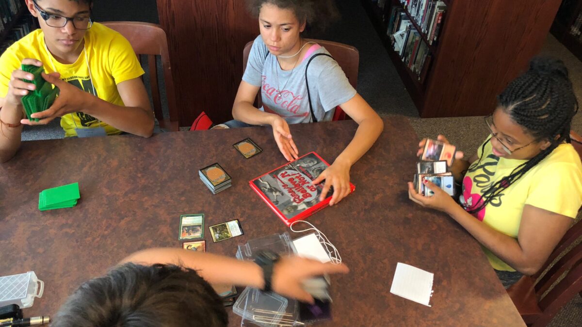 Three youth interacting with trading cards and a red book on a brown table