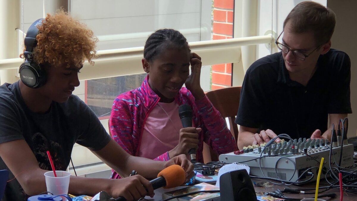 Adult in front of a mixing board looking at two youth holding microphones, one wearing headphones