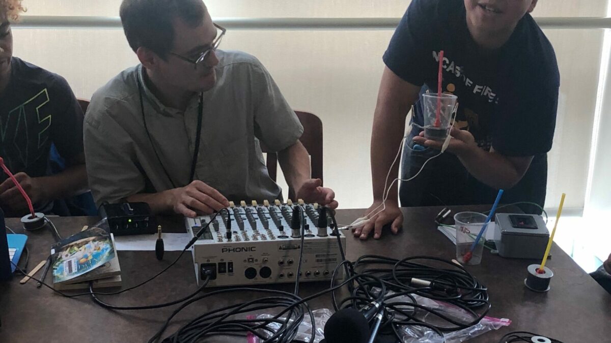 Youth holding a plastic cup, as an adult in front of a mixing board looks on