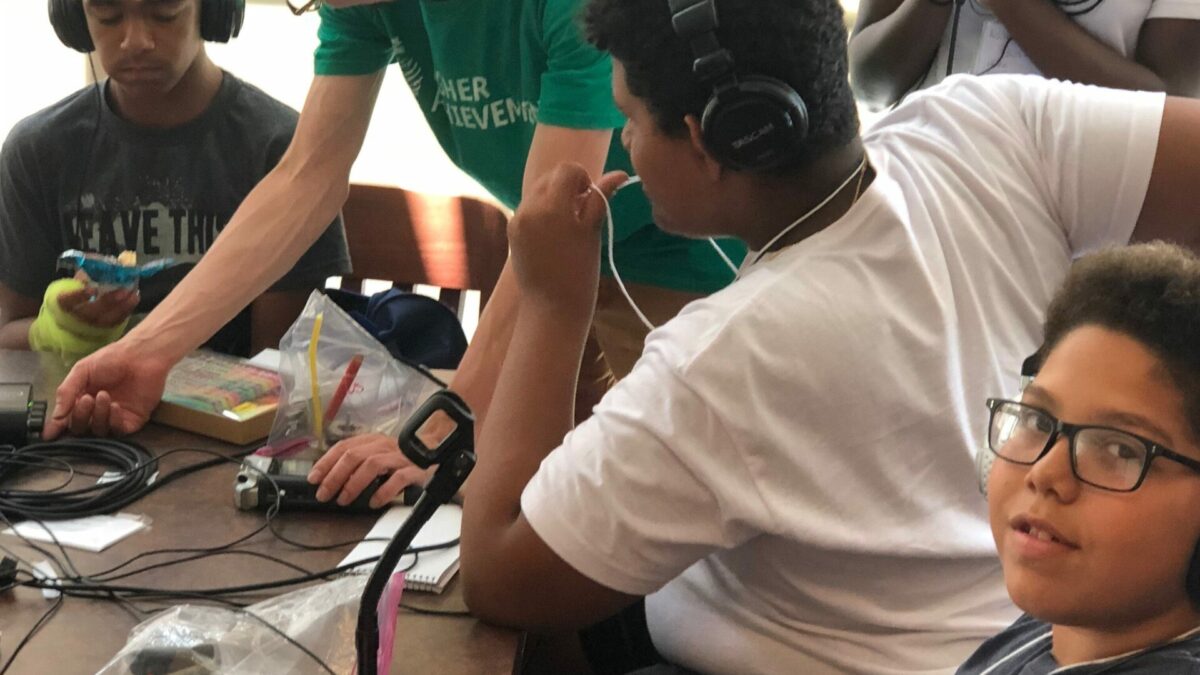 Four youth and an adult in front of a brown table, two youths wearing headphones, one youth holding and speaking into a microphone