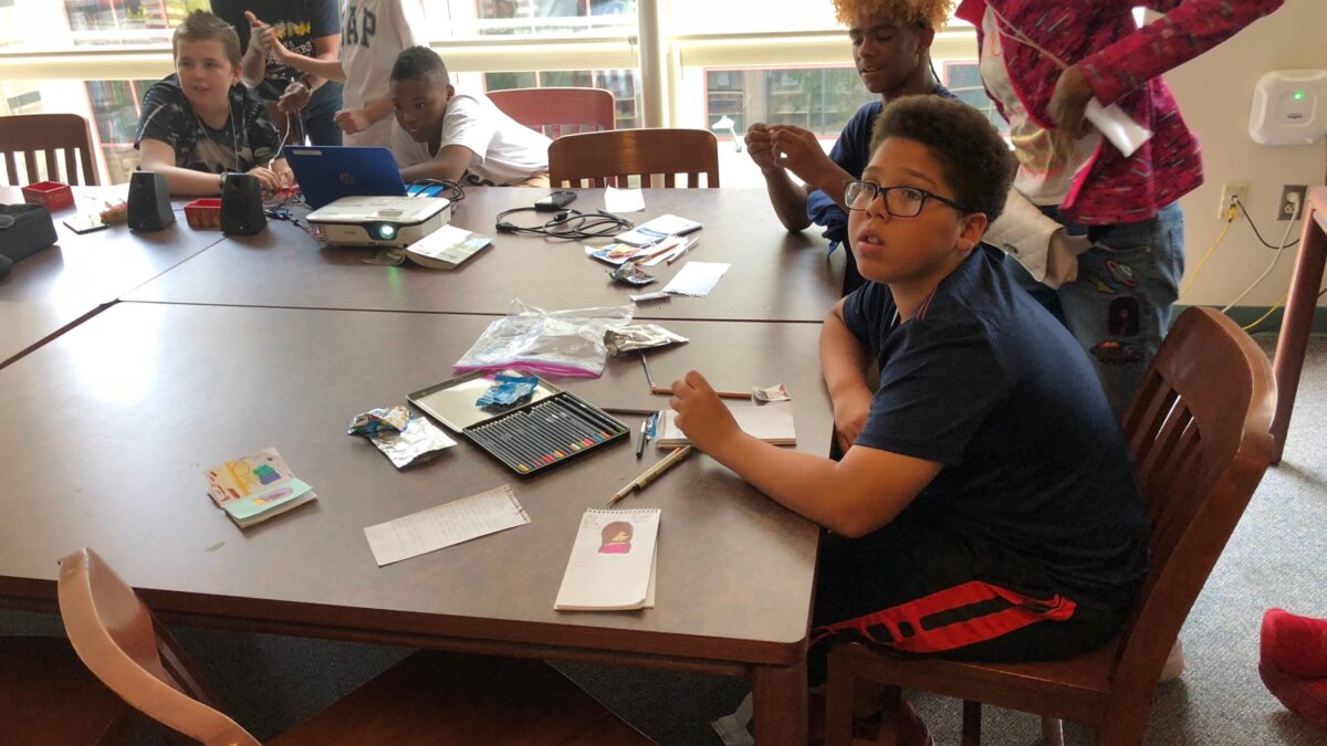 Six youth and one adult around multiple brown tables, the adult and three youth in front of a blue laptop