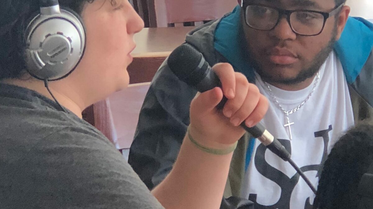 Teen wearing headphones speaking into a microphone as another teen watches