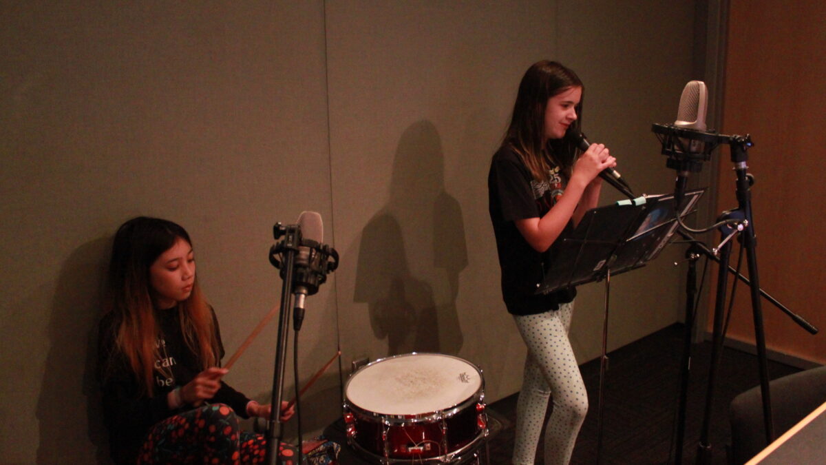 Two youth, one playing drum set, the other singing into a microphone and looking at sheet music on a music stand in a radio studio