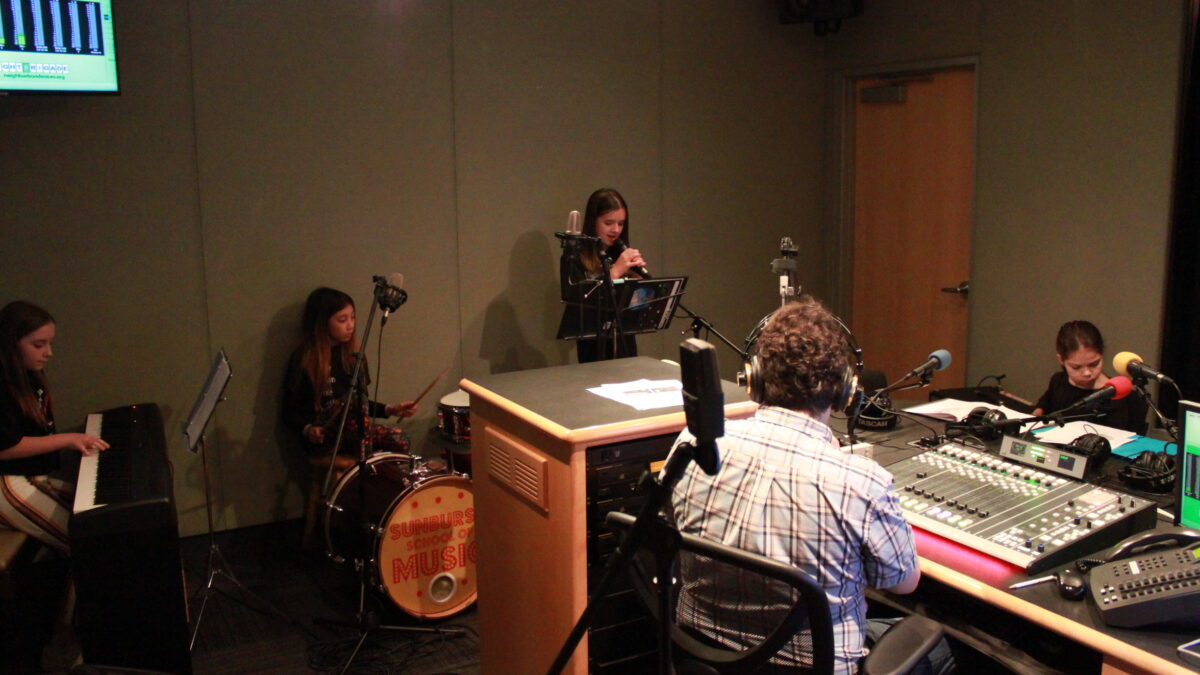 Four youth musicians performing on guitar, piano keyboard, drum set, and voice for an adult wearing headphones in a radio studio