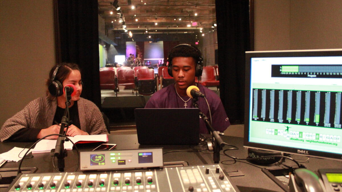 Two teens wearing headphones and looking at a laptop in front of microphones in a radio studio