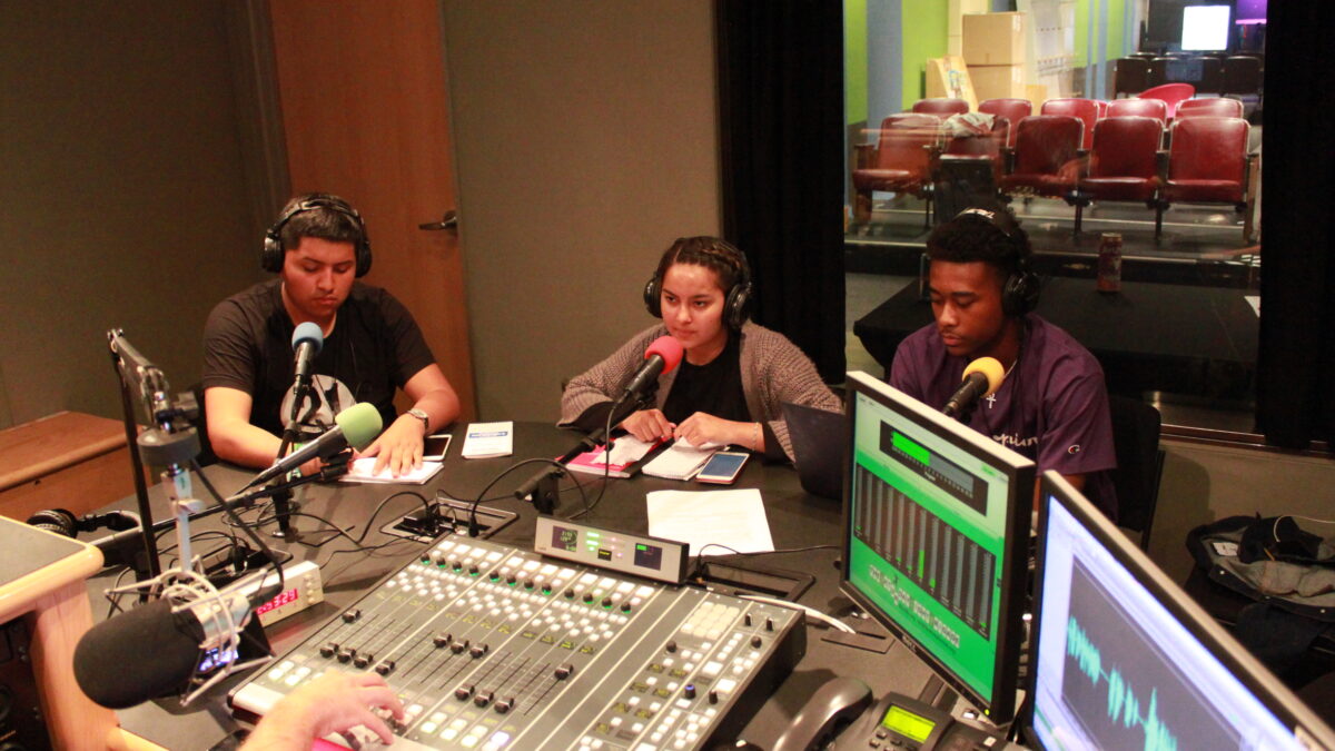 Three teens wearing headphones sitting in front of microphones being interviewed in a radio studio