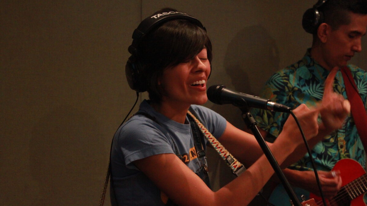 Young adult wearing headphones, holding a ukulele and singing into a microphone in a radio studio
