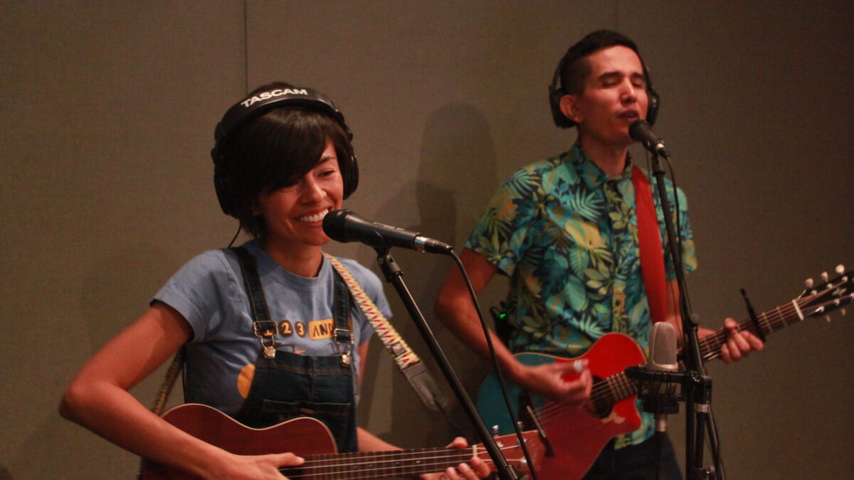 Two young adults wearing headphones and singing into microphones in a radio studio, one playing ukulele, the other playing guitar