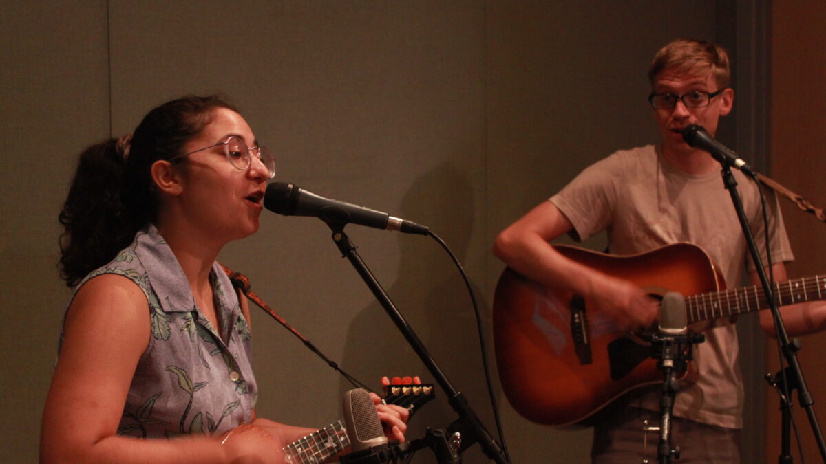 Two young adults singing into microphones in a radio studio, one playing ukulele, the other playing guitar