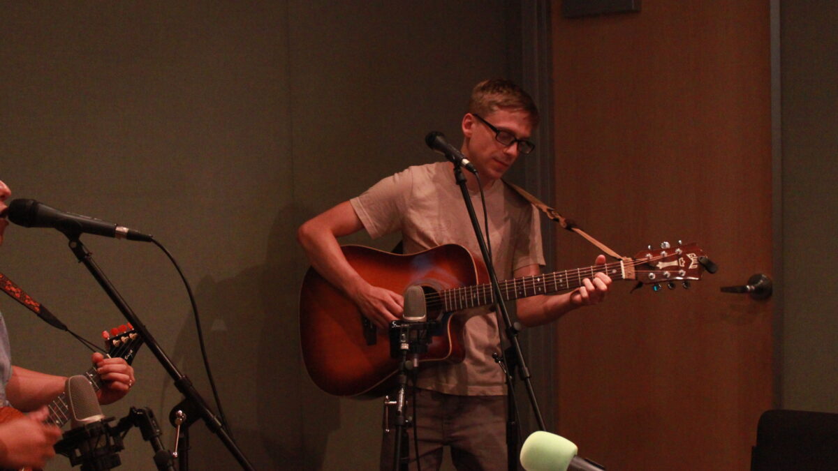 Adult playing guitar in front of a microphone in a radio studio