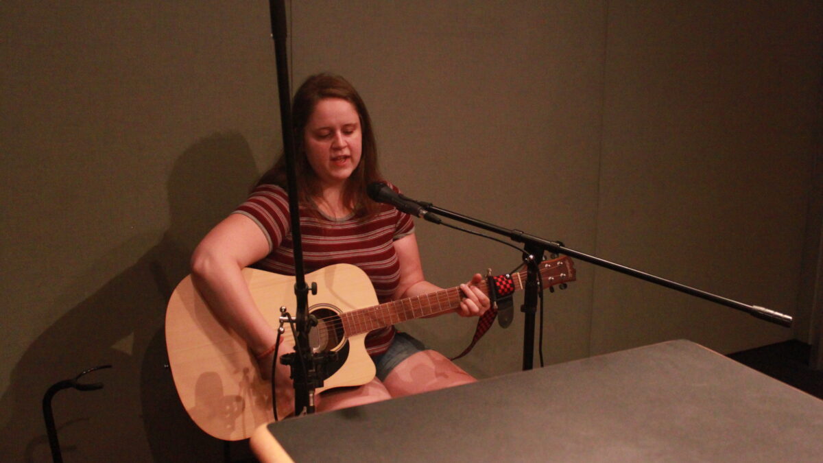 Adult playing guitar and singing into a microphone in a radio studio