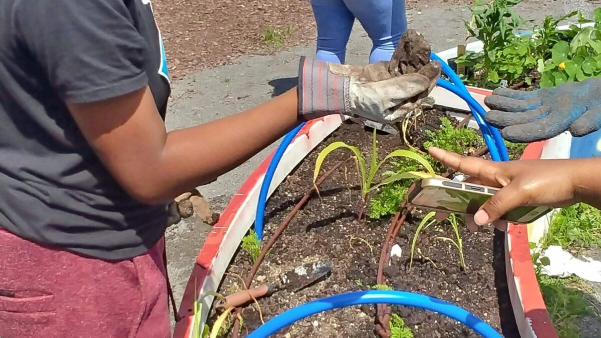 One youth holding a small mound of dirt and smiling, with another youth holding a large blue watering can watering plants in a planter