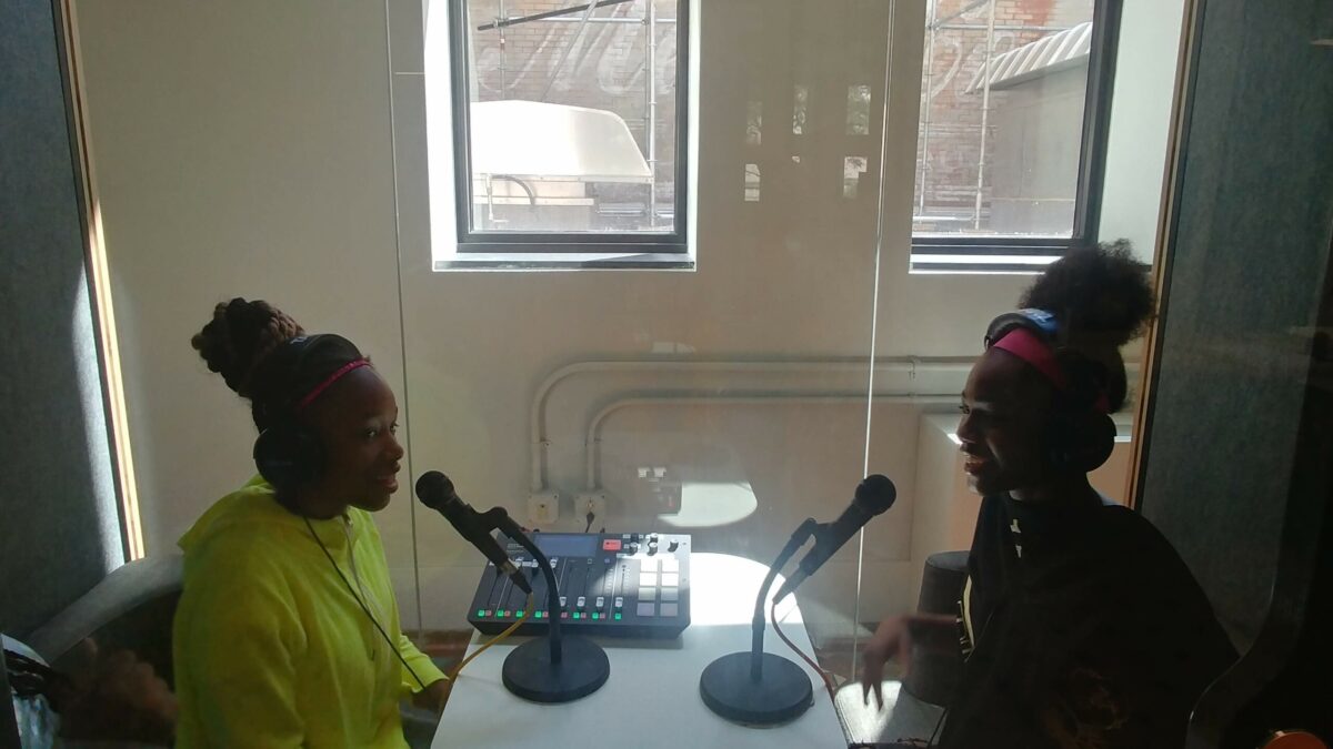 Two youth wearing headphones and speaking into microphones in a small recording pod with a mixing board in the middle